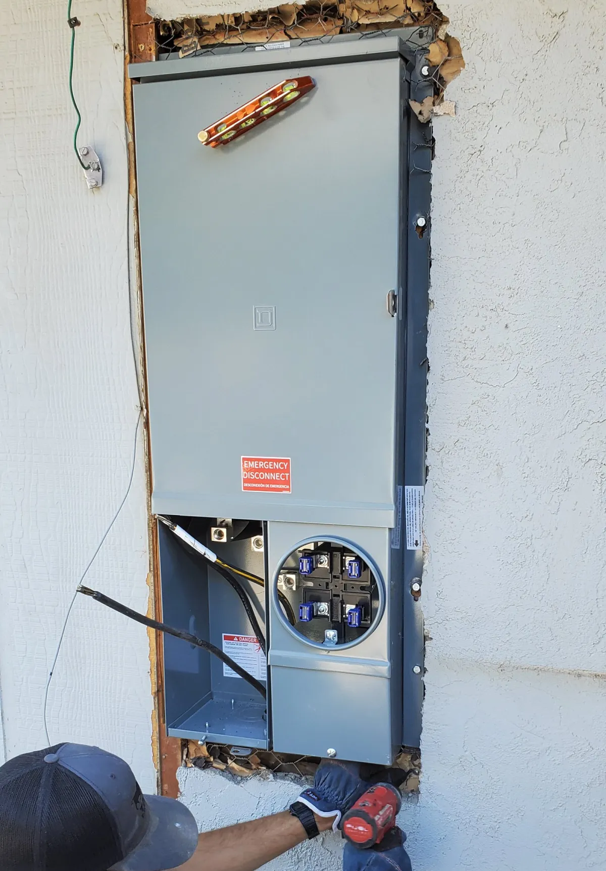 A Taurus electrician fastening a new grey meter main panel into a freshly cut opening in a stucco wall